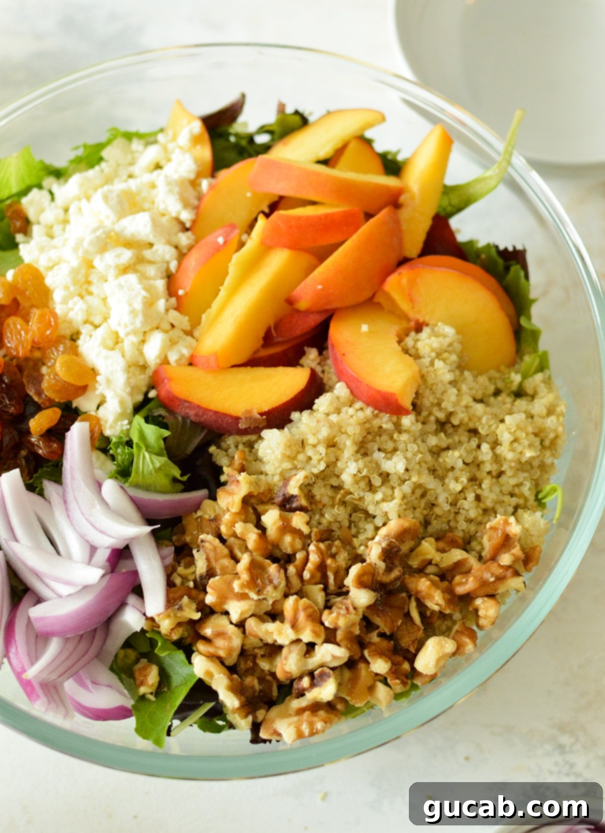 Peachy Feta Quinoa Salad 4 Unmixed quinoa salad ingredients neatly arranged in a large bowl: featuring vibrant peaches, fluffy quinoa, crunchy walnuts, creamy feta, and sweet raisins atop a bed of mixed greens, awaiting the dressing.