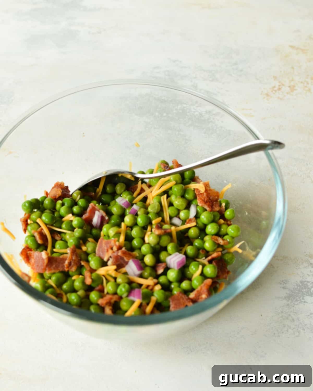 A large bowl containing the main salad ingredients: bright green peas, shredded cheddar cheese, diced red onion, and crumbled crispy bacon, before the dressing is added.