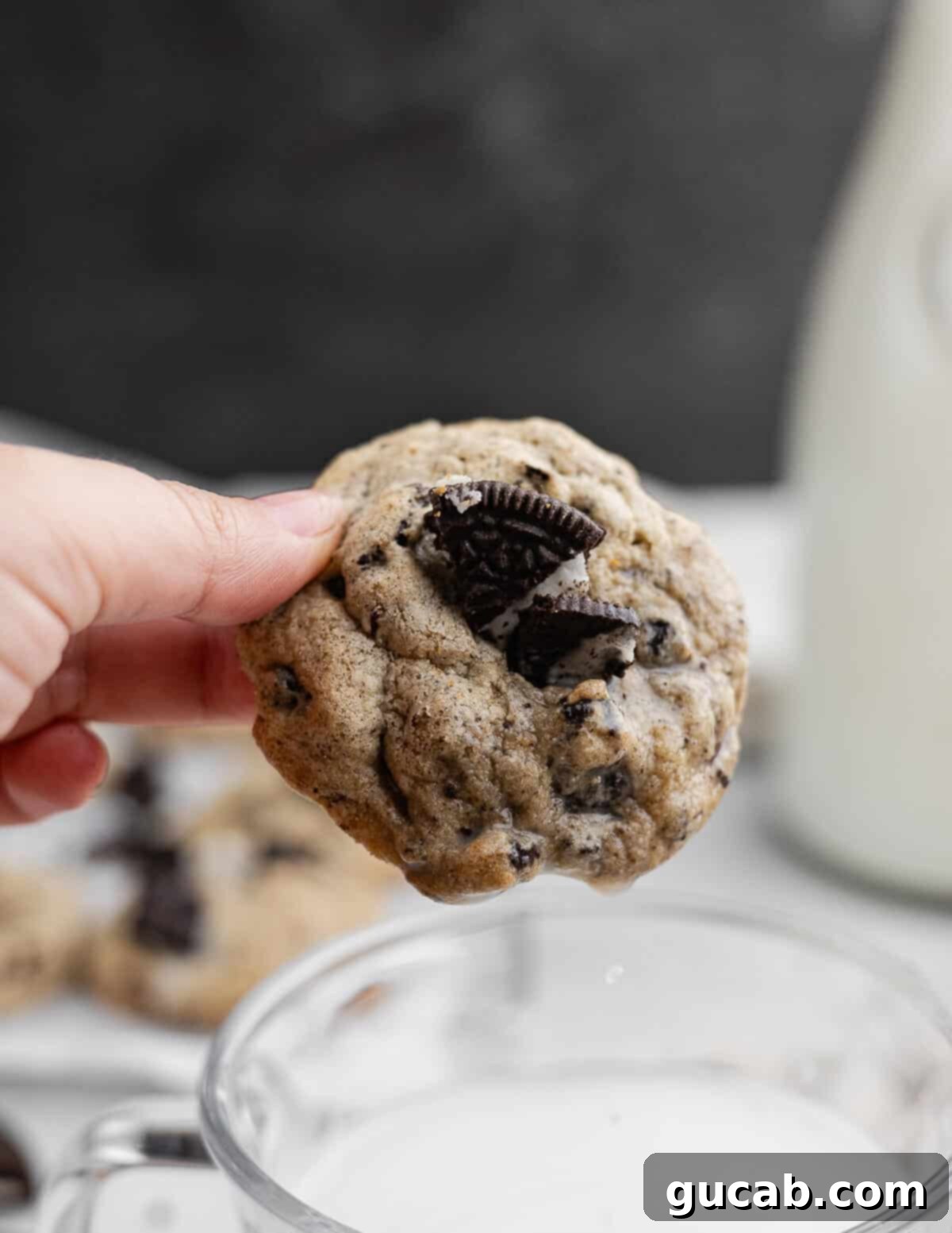 A hand gently holding a freshly baked Oreo chocolate chip cookie, poised above a frosty glass of milk, highlighting its golden edges, soft texture, and visible Oreo pieces.