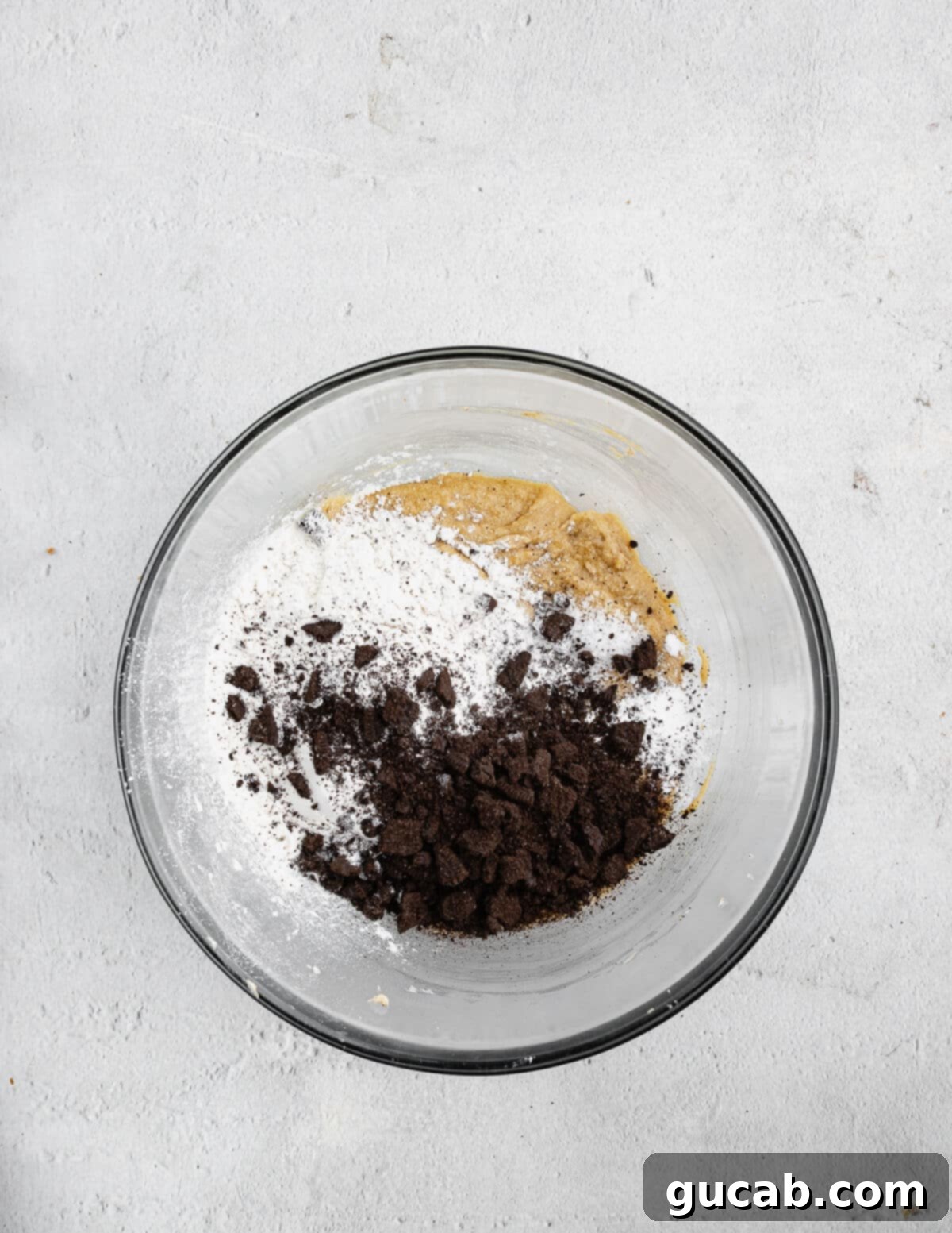 A mixing bowl containing creamed butter, sugars, and eggs, with the dry ingredients – flour, baking soda, salt, and Oreo cookie crumbs – carefully layered on top, ready for mixing.