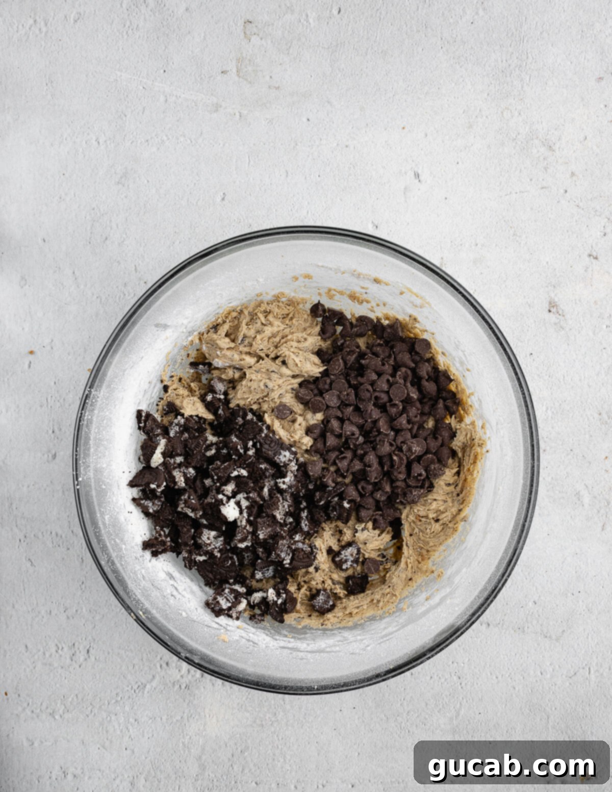 A mixing bowl filled with cookie dough, topped with generous amounts of roughly broken Oreo pieces and semi-sweet chocolate chips, ready to be folded in.