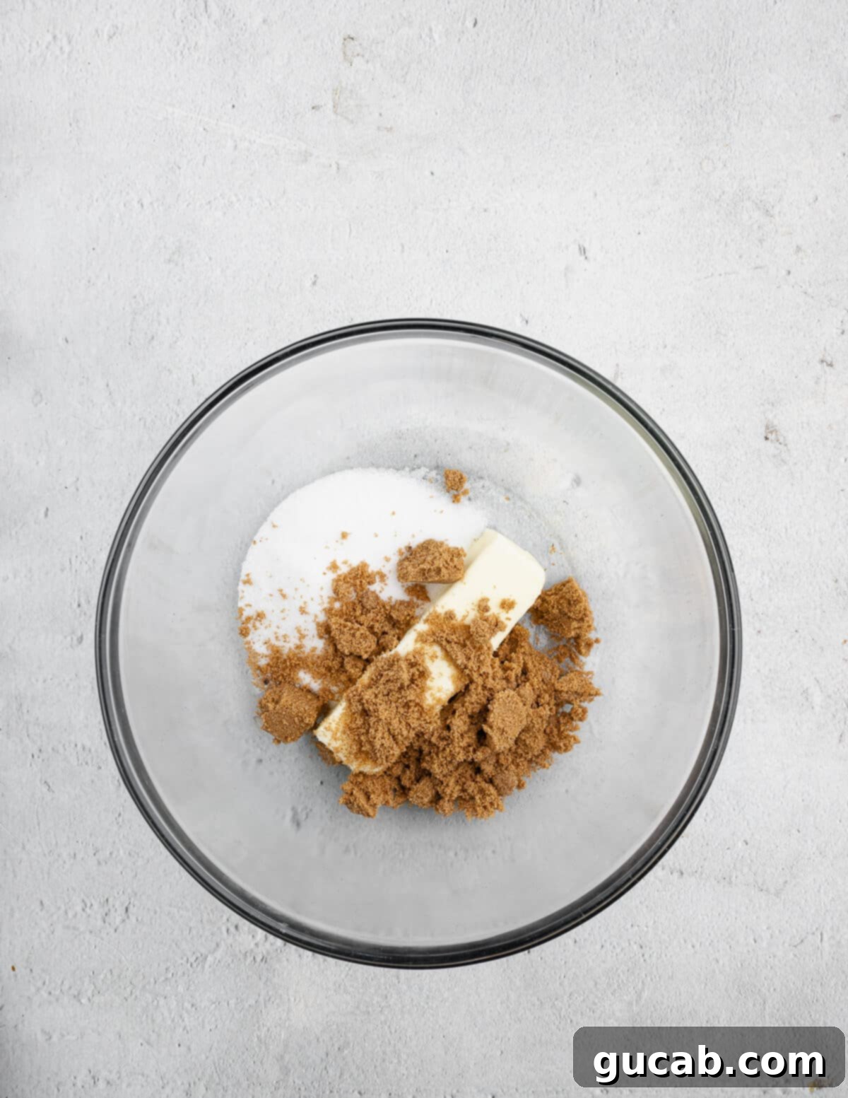 A stick of unsalted butter, granulated sugar, and brown sugar are placed in a mixing bowl, ready to be creamed together for the cookie dough.