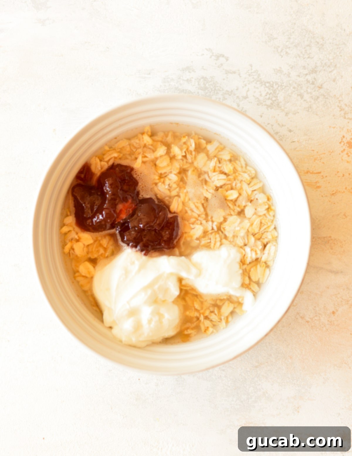 A bowl containing rolled oats, water, Greek yogurt, raspberry jam, and maple syrup, ready to be mixed for overnight oats.