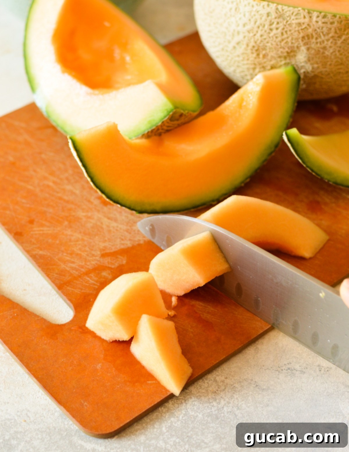 A knife actively cutting a cantaloupe slice into smaller chunks on a cutting board.