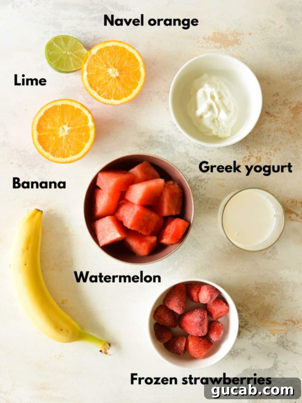 A selection of fresh ingredients including watermelon chunks, bananas, oranges, limes, frozen strawberries, and Greek yogurt, arranged for a watermelon banana smoothie.