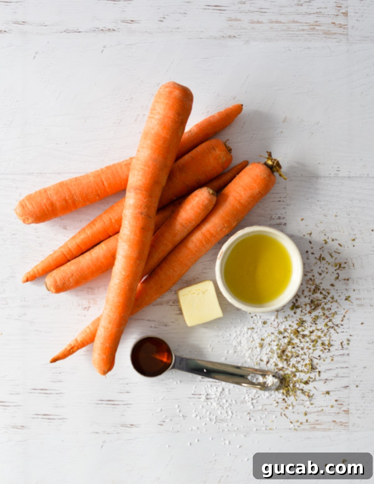 a flat lay of all the essential ingredients for maple glazed carrots: fresh whole carrots, a bottle of olive oil, a stick of unsalted butter, and a bottle of rich maple syrup, all arranged on a rustic wooden surface.