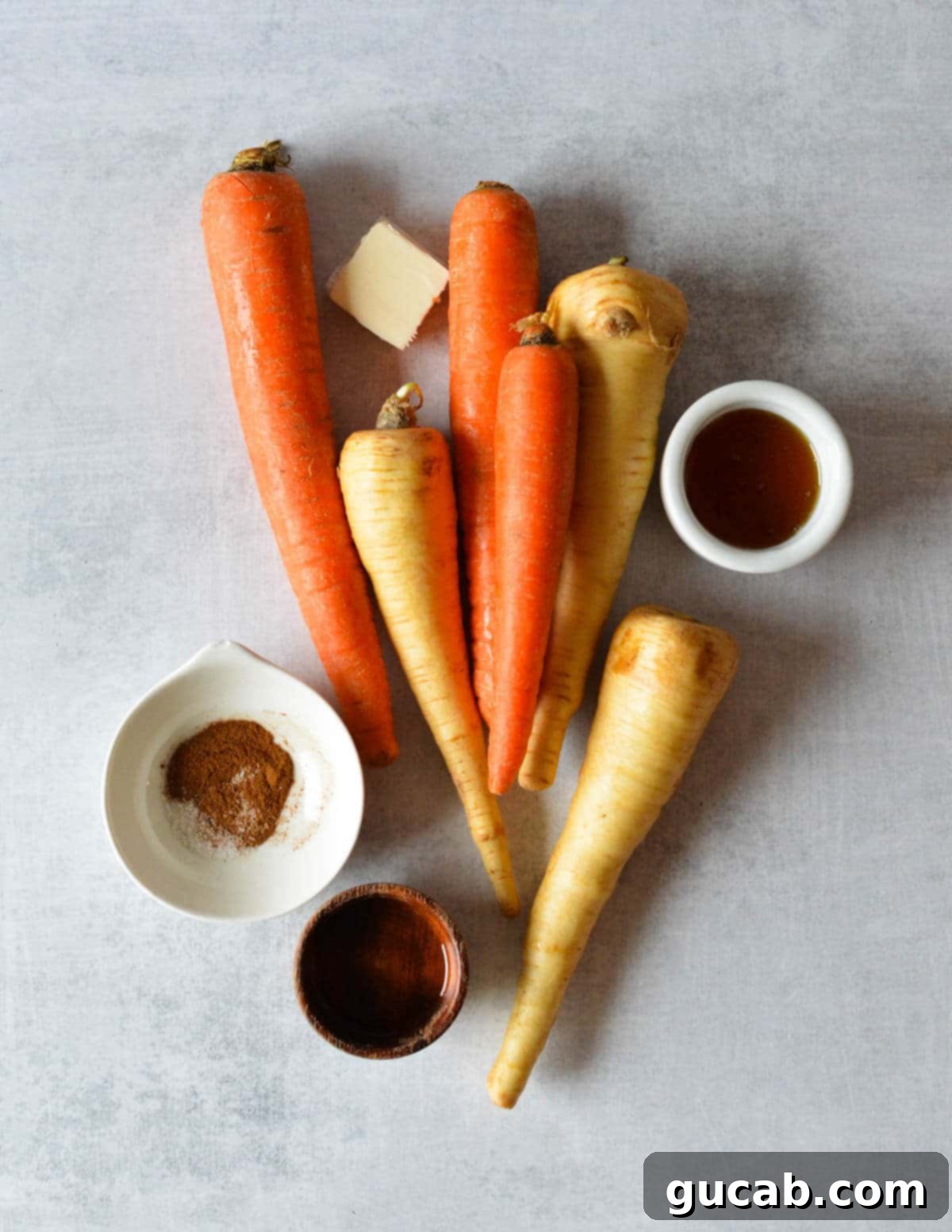 A flat lay photo showing all the key ingredients: fresh carrots, parsnips, a bottle of olive oil, a stick of butter, a jar of honey, and small bowls of cinnamon and salt.