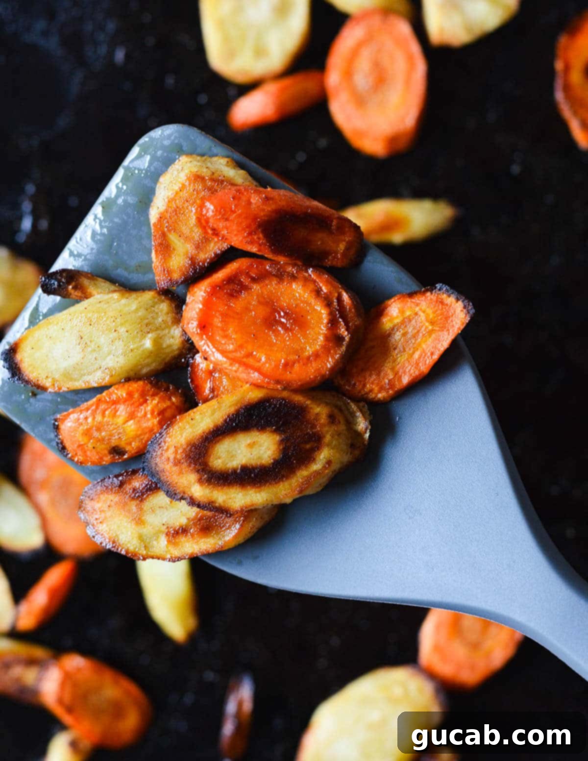 Close up of perfectly roasted carrots and parsnips being lifted off a sheet pan with a rubber spatula, showing their golden-brown caramelization.