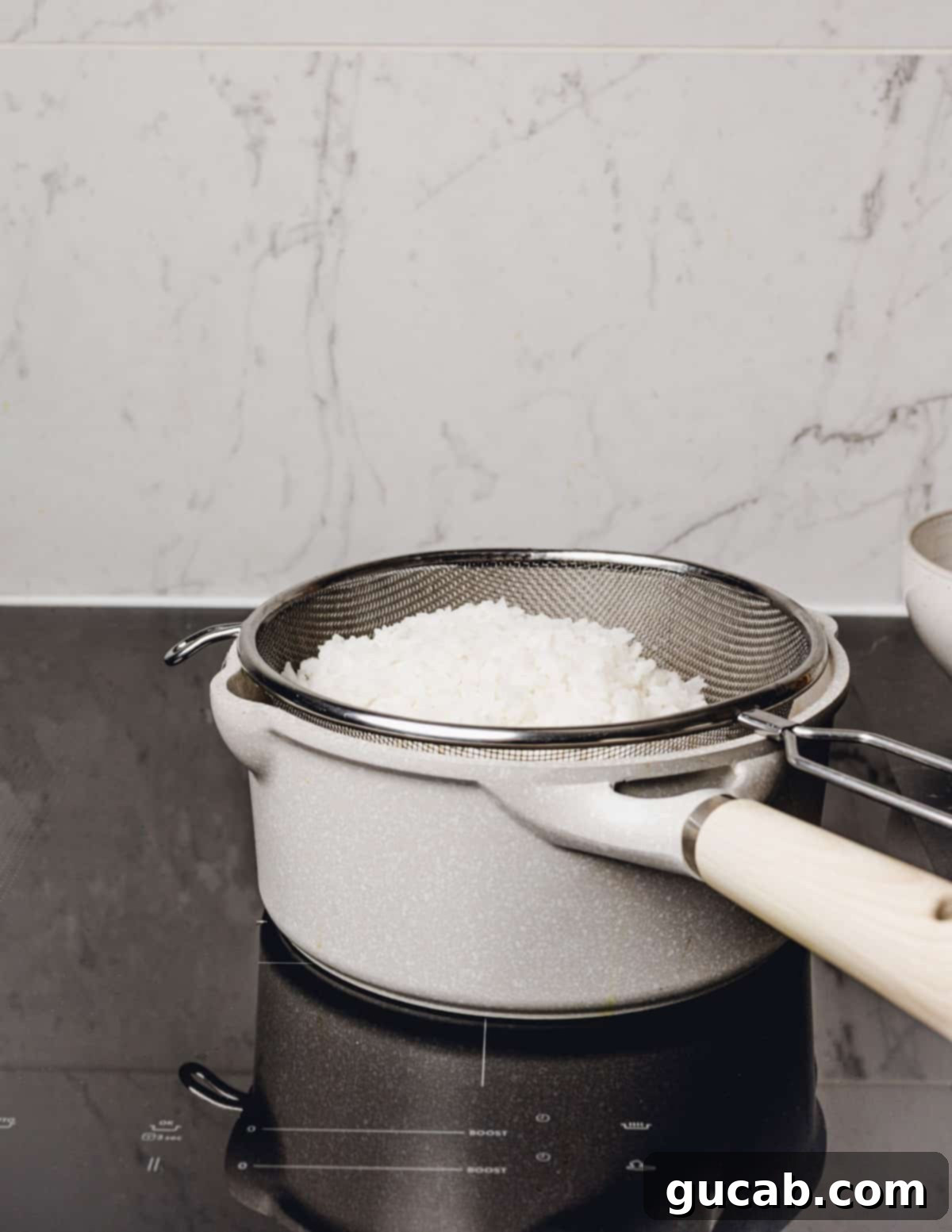 A strainer full of cooked white rice set on top of the saucepan, allowing residual steam to escape and rice to cool slightly.