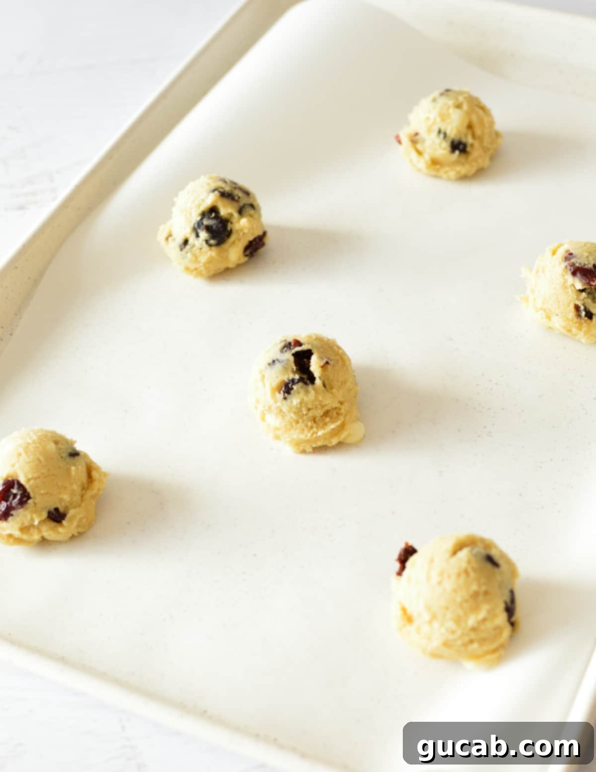 Six evenly scooped cookie dough balls with visible dried cherries and white chocolate chips, arranged on a baking sheet lined with parchment paper.