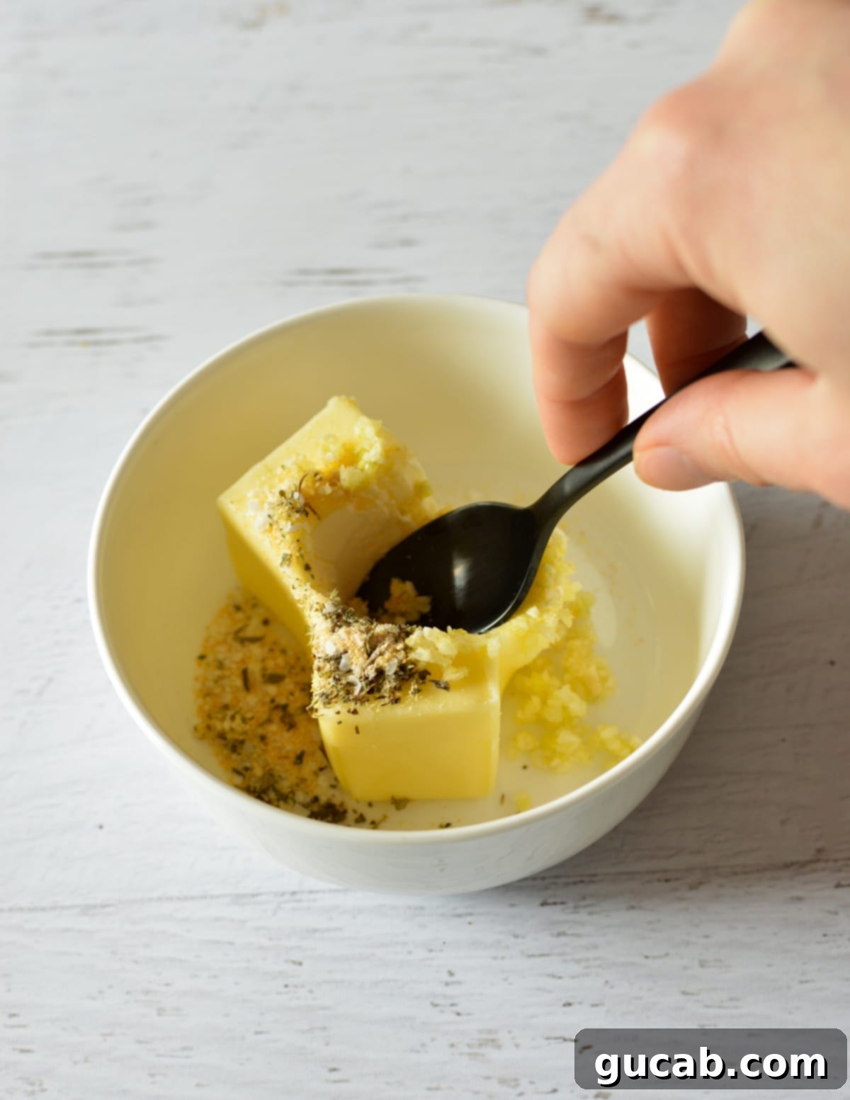 A spoon mixing softened butter with minced garlic and herbs in a white bowl.