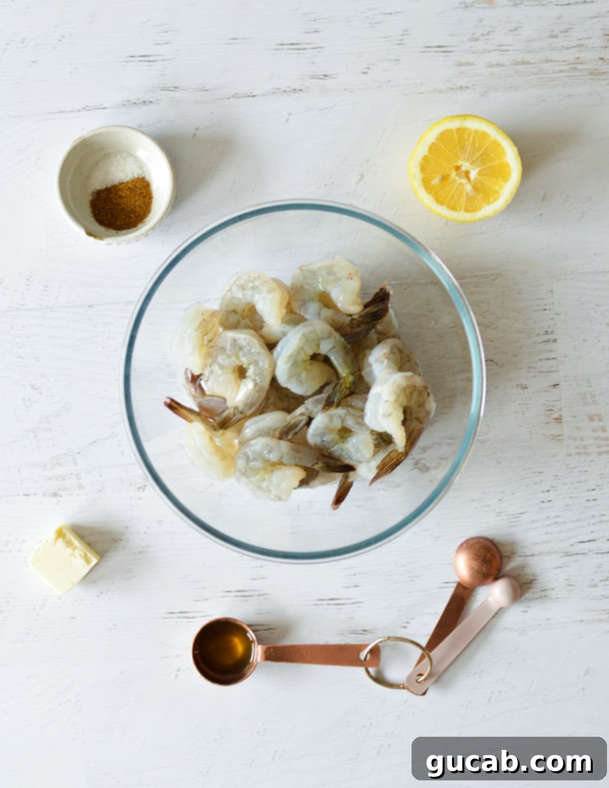 Fresh shrimp, butter, olive oil, Old Bay seasoning, and lemon slices laid out on a surface.