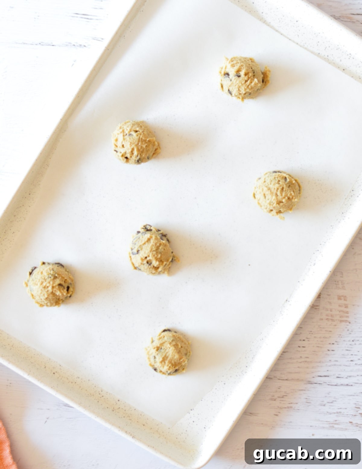 Six evenly spaced chocolate chip cookie dough balls on a parchment-lined baking sheet, topped with extra chocolate chips.