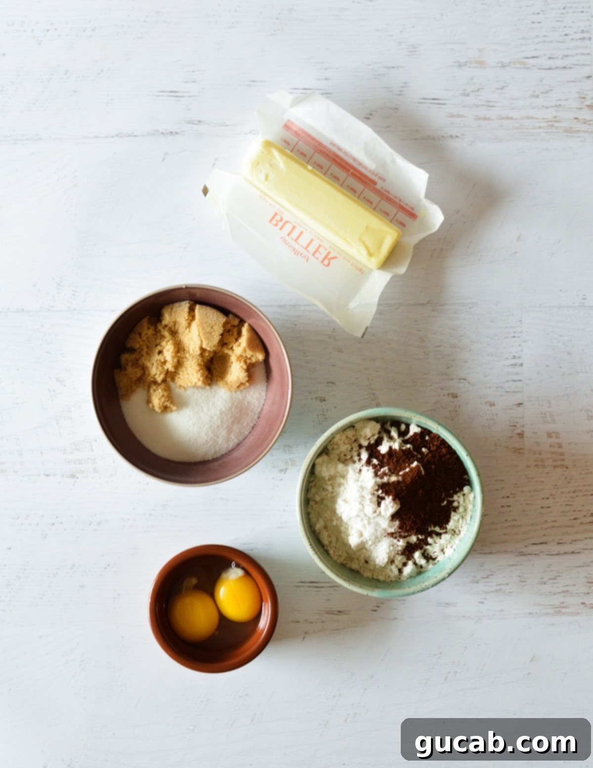 A selection of coffee cookie ingredients: butter, sugar, brown sugar, flour, instant coffee, and eggs, laid out on a surface.