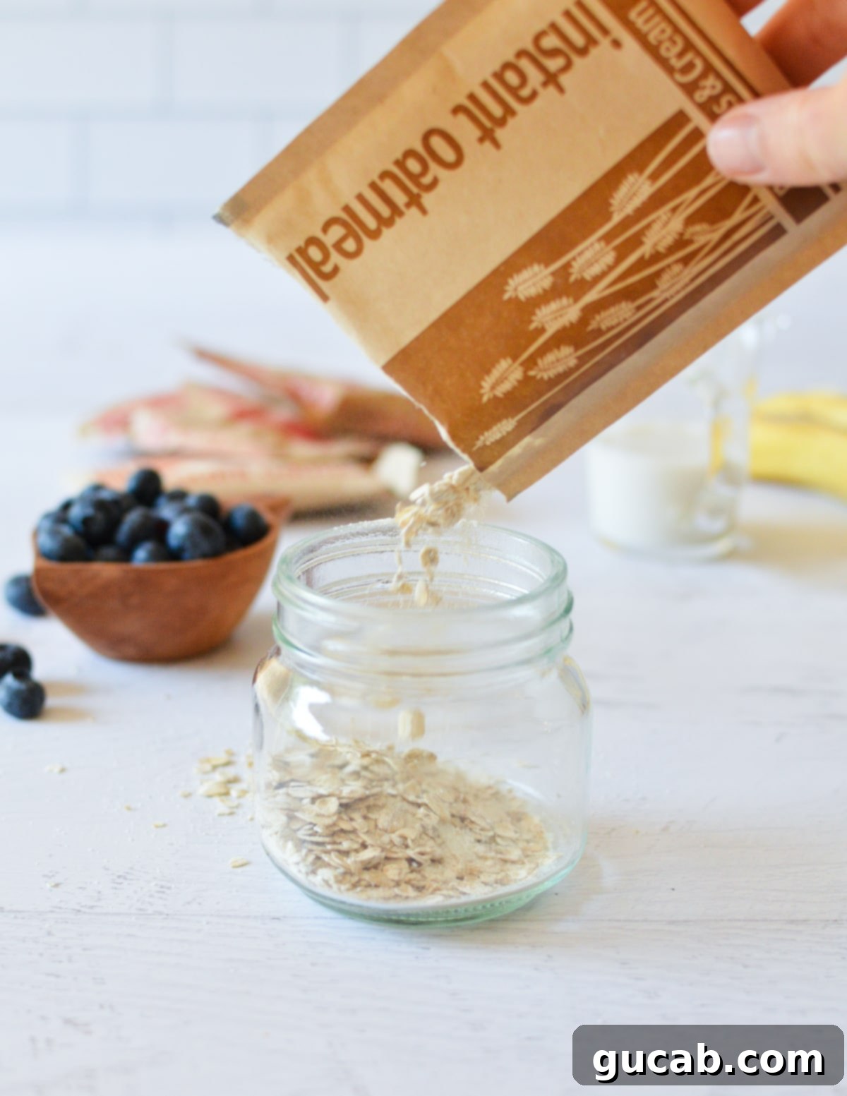 A hand gently emptying an instant oatmeal packet into a clear glass jar, showing the dry oats settling at the bottom.
