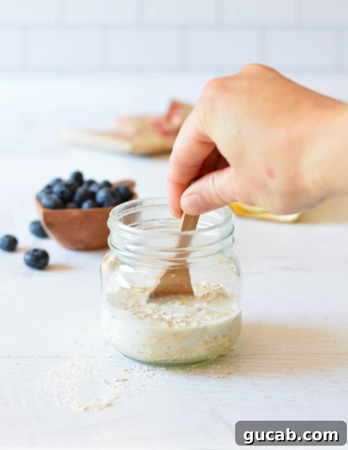 A close-up of a hand stirring cold oats and milk in a jar with a spoon, ensuring all ingredients are fully combined.