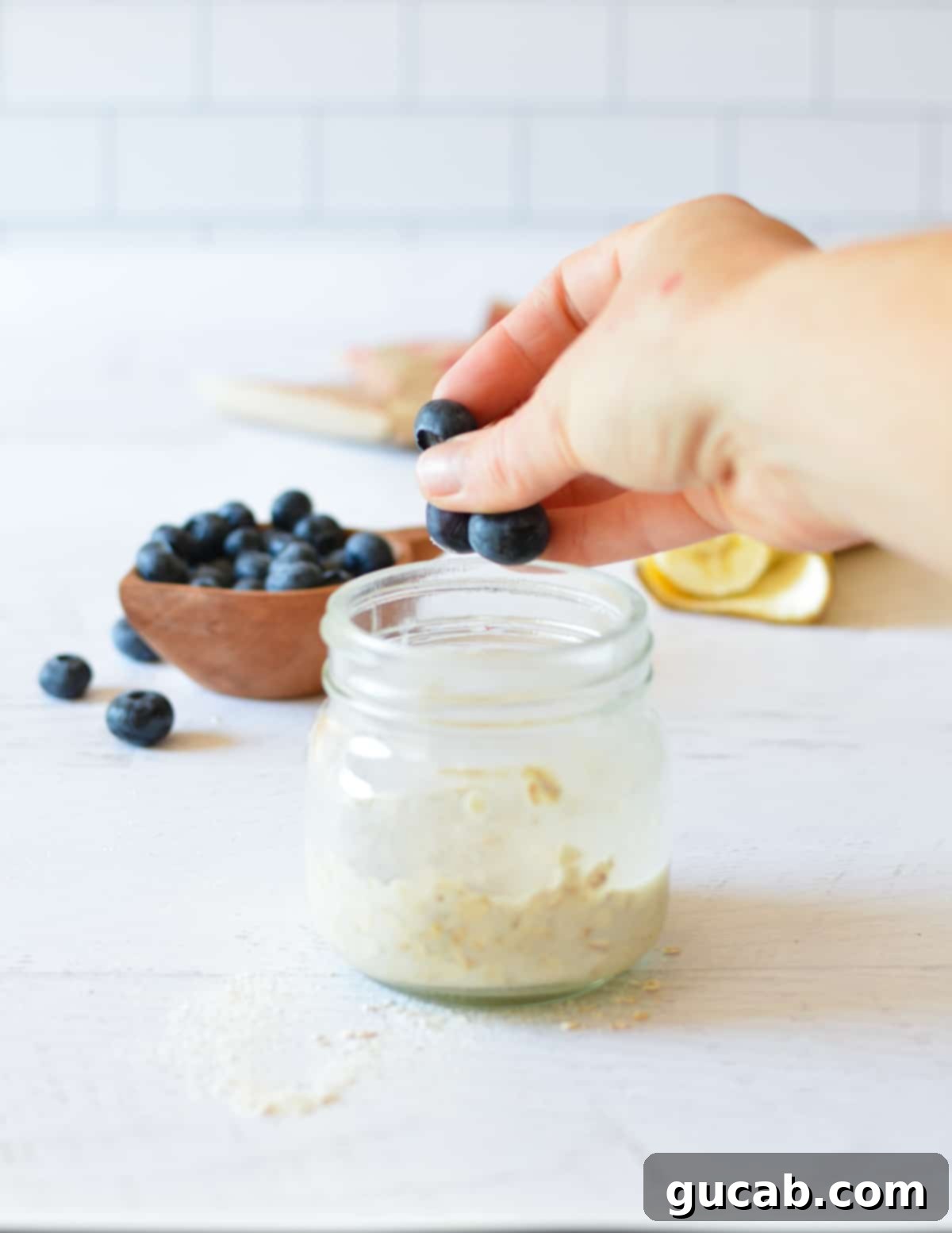 A hand gently dropping fresh blueberries into a jar of mixed overnight oats, adding a burst of color and flavor.