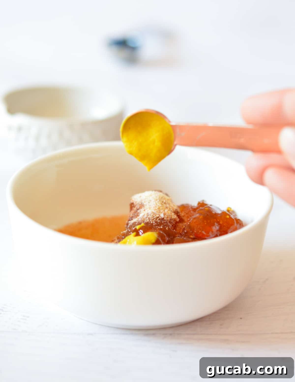 A hand pouring yellow mustard into a bowl containing apricot jam, spices, and pineapple juice, ready to be mixed.