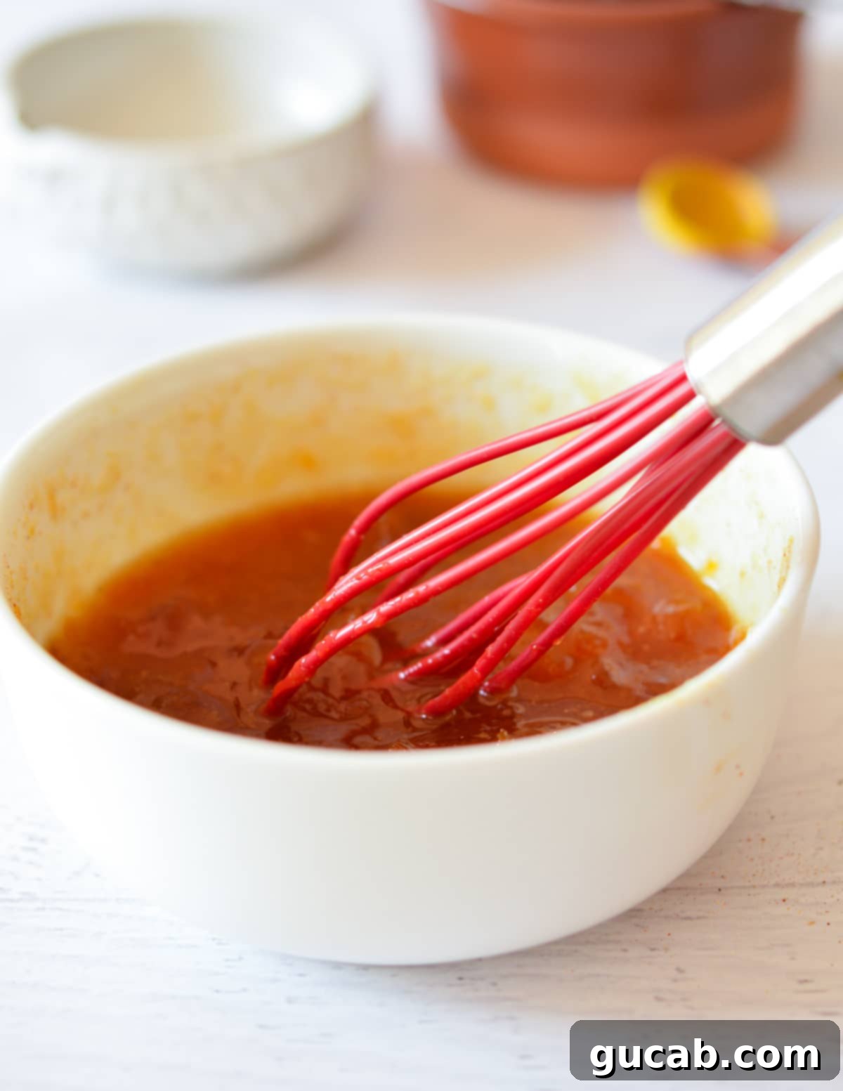 A close-up of a whisk stirring the sweet and sour sauce in a clear glass bowl, showing its smooth consistency.