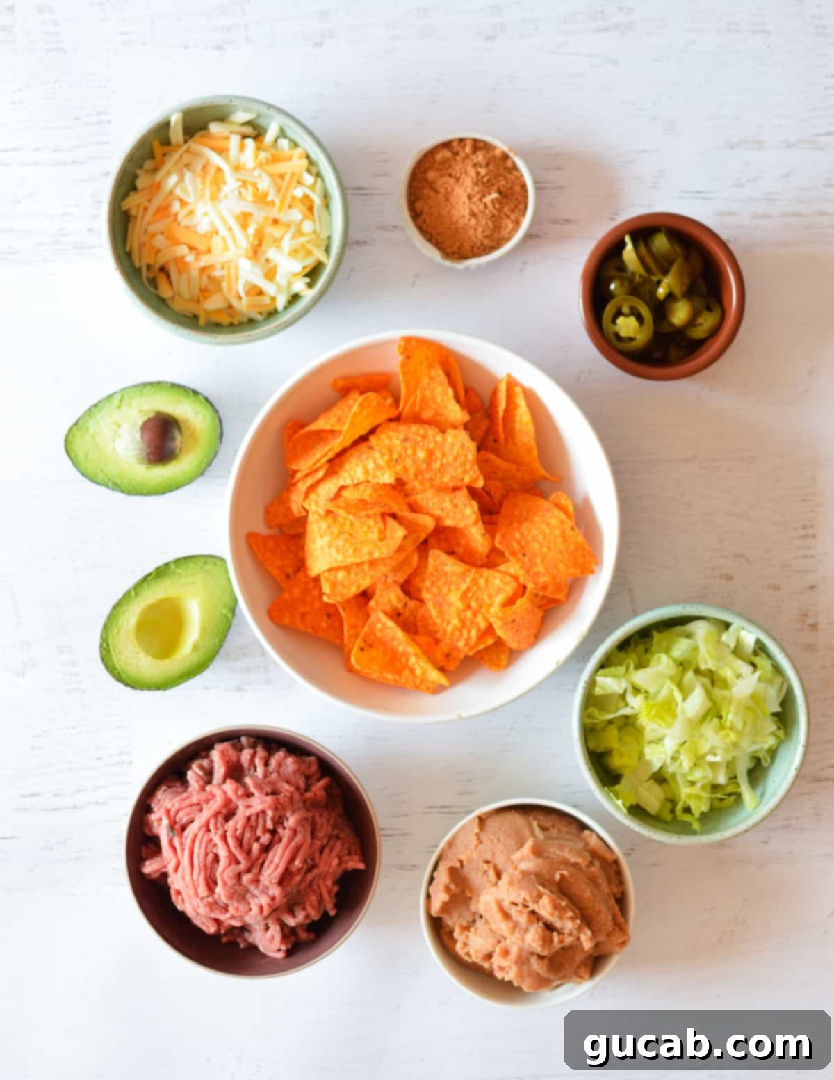 A flat lay of key ingredients for Doritos nachos: Doritos, cheese, taco seasoning, pickled jalapenos, avocado, lettuce, ground beef, and refried beans.