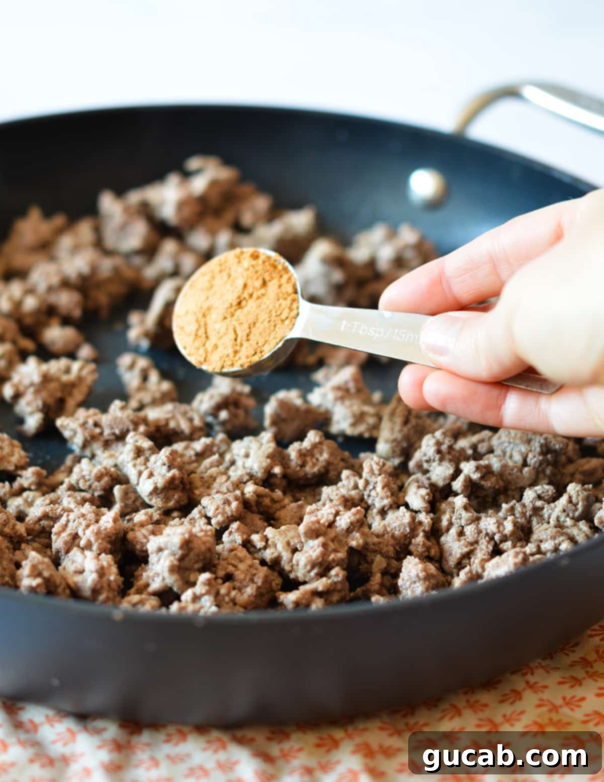 A hand sprinkling taco seasoning over browned ground beef in a skillet.