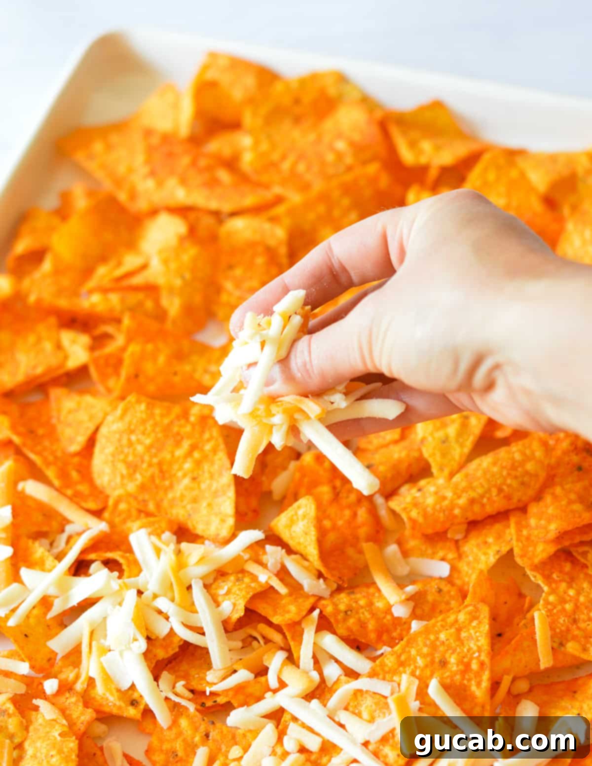 A hand sprinkling shredded cheese generously over Doritos chips on a baking sheet.