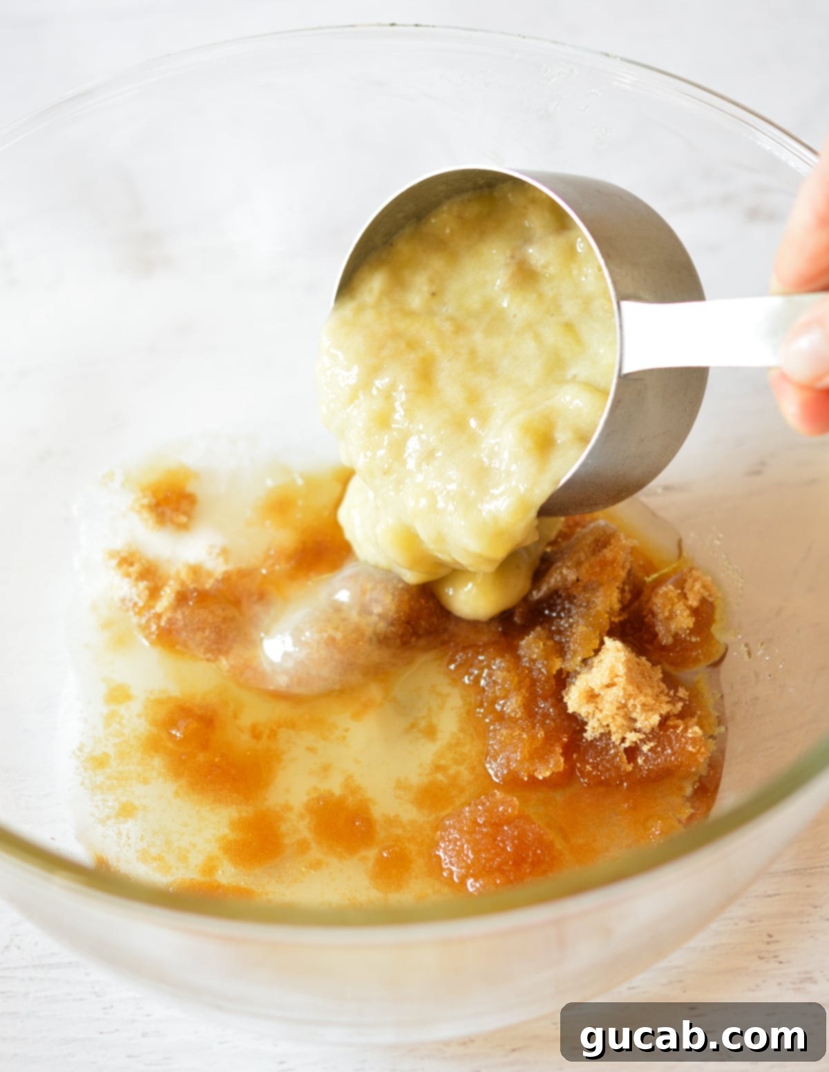 The process of pouring mashed banana into a large bowl containing white and brown sugar, alongside oil and vanilla.
