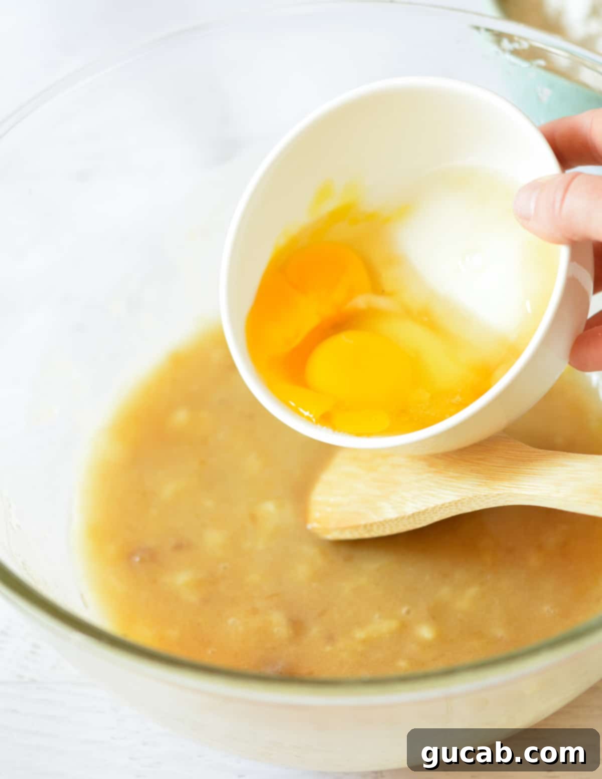 Cracked eggs being poured into the bowl of mashed bananas, sugar, and oil, ready to be mixed.