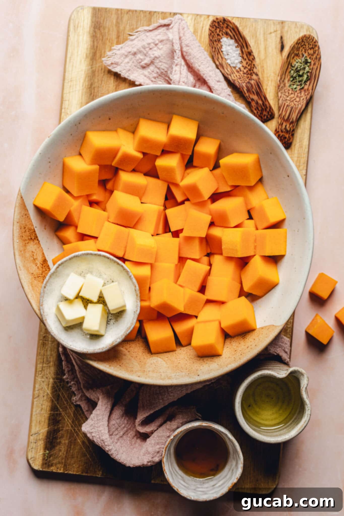 A visually appealing flat lay of the essential ingredients for sauteed butternut squash: neatly cubed butternut squash, a small bowl of olive oil, a stick of unsalted butter, a pour of maple syrup, a pinch of kosher salt, and a sprinkle of dried thyme.