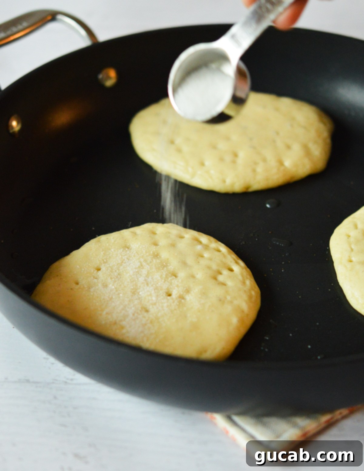 A hand carefully sprinkling granulated sugar onto the cooking pancakes in a hot skillet, preparing them for flipping.