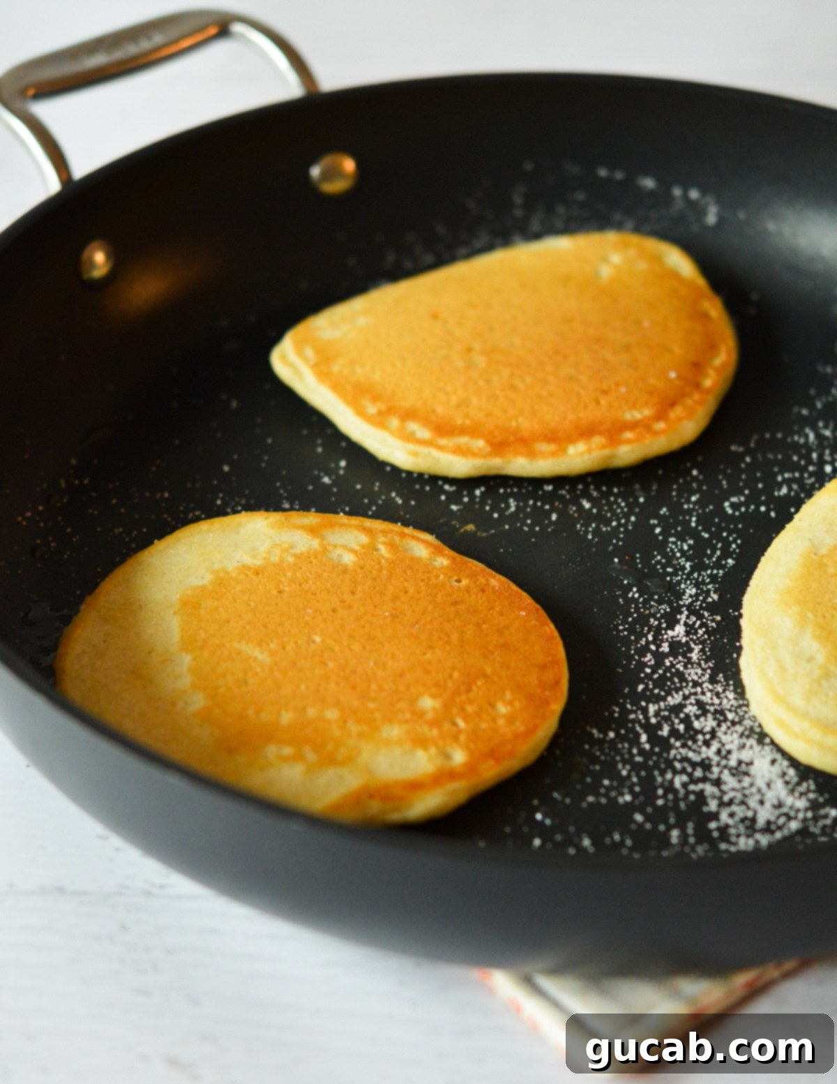 Crispy pancakes cooking in a skillet after being flipped, showing golden-brown bottoms achieved from caramelized sugar.