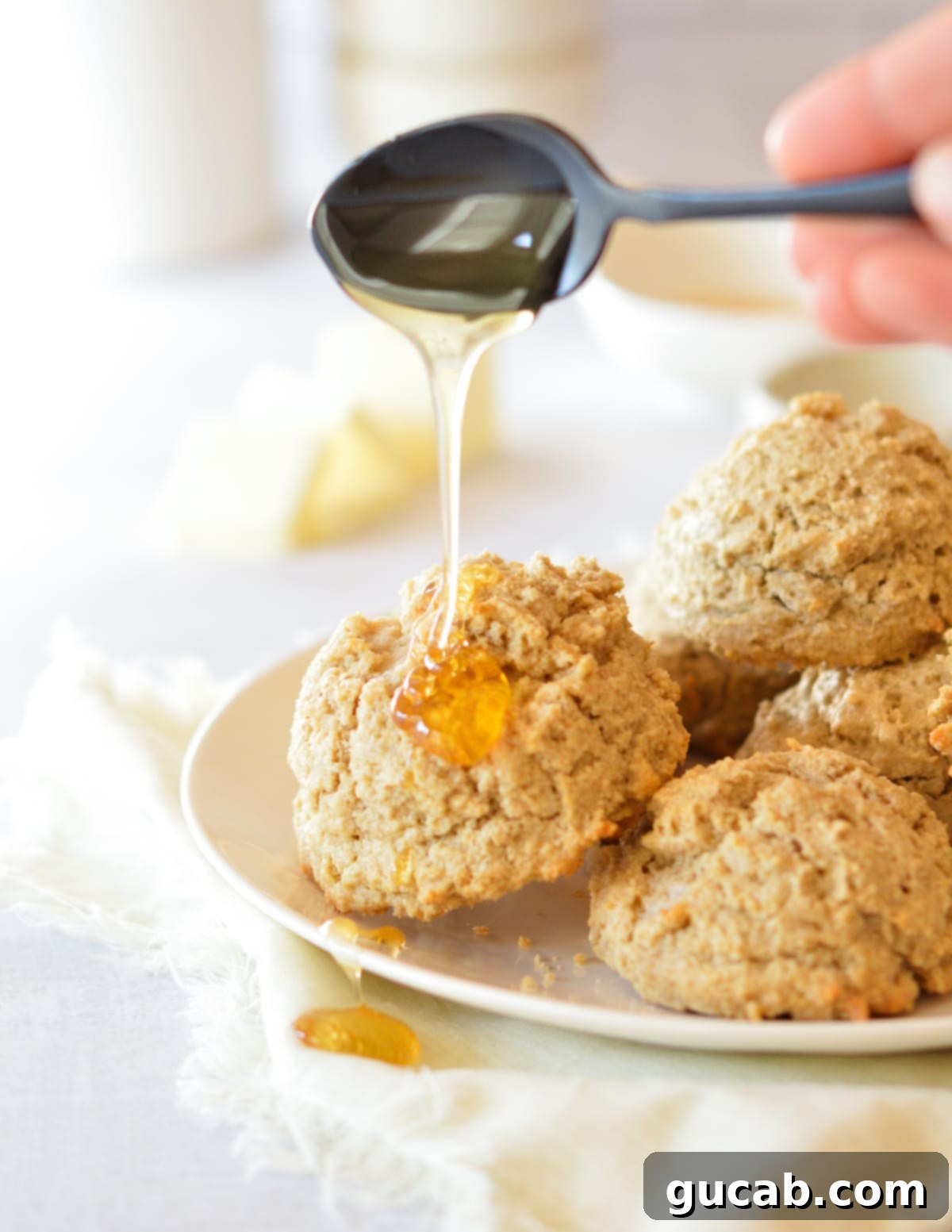 A stream of golden honey being generously poured over a stack of warm, golden brown oat flour biscuits on a white plate.