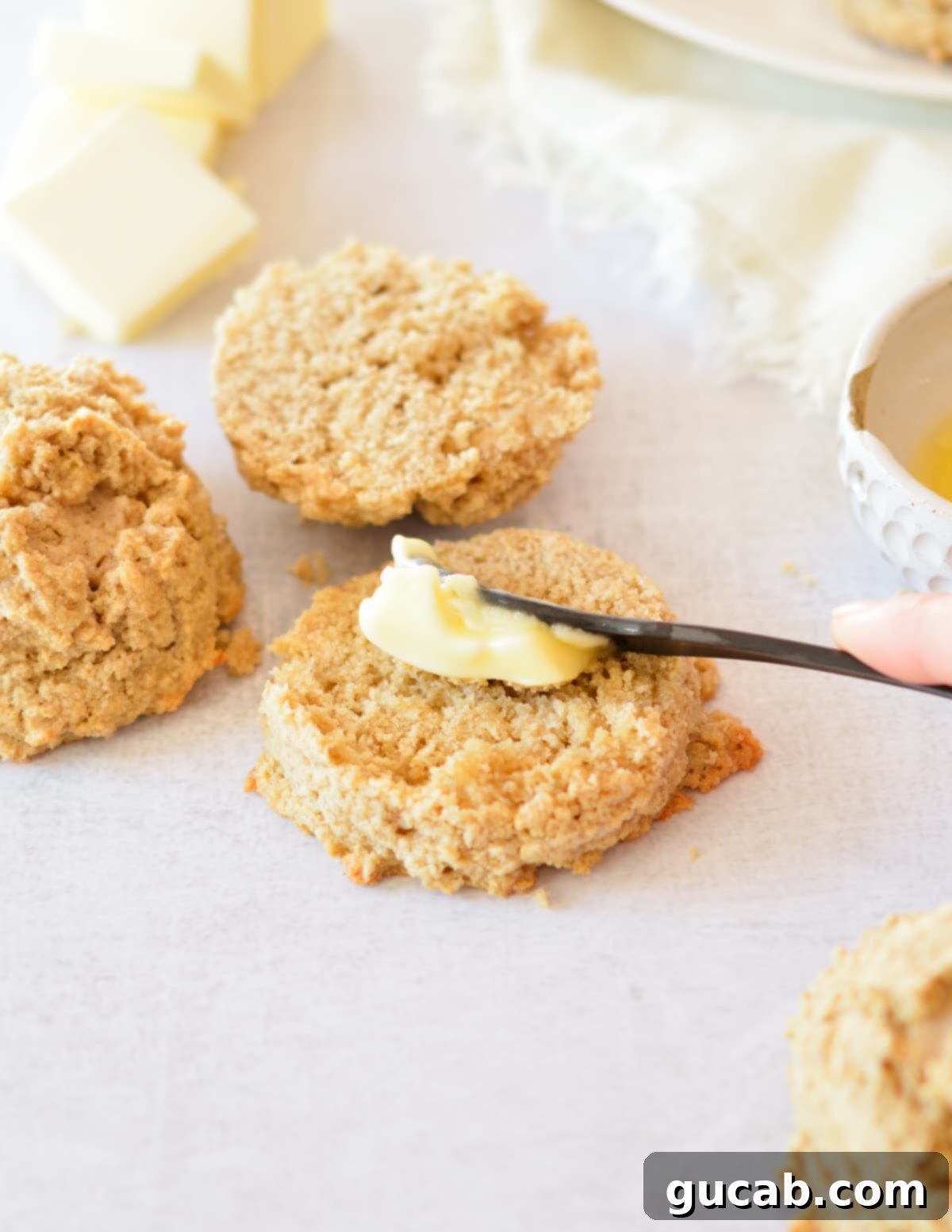 A knife is gently spreading creamy butter onto the fluffy, golden interior of a halved oat flour biscuit.