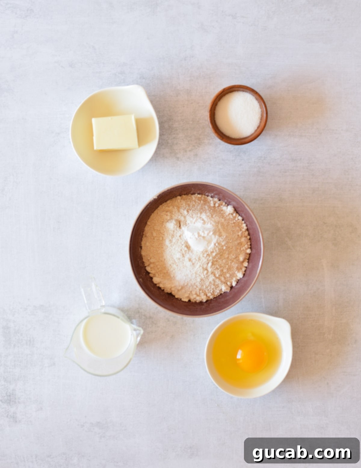 A flat lay photograph showcasing the key ingredients for oat flour biscuits: cold butter, granulated sugar, fine oat flour, a fresh egg, and a bottle of milk.