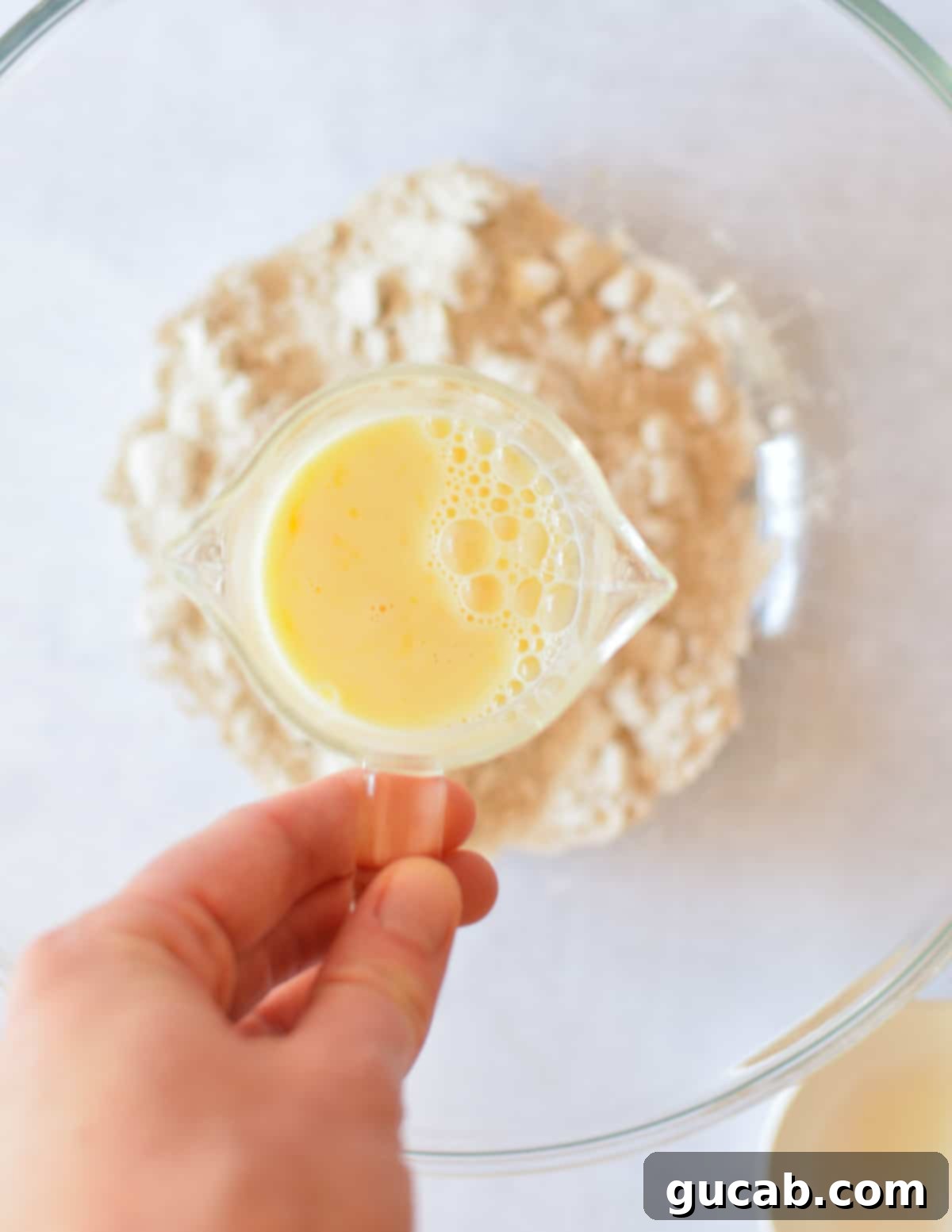 A hand holds a measuring cup containing milk and a whisked egg, ready to be added to the dry ingredients.