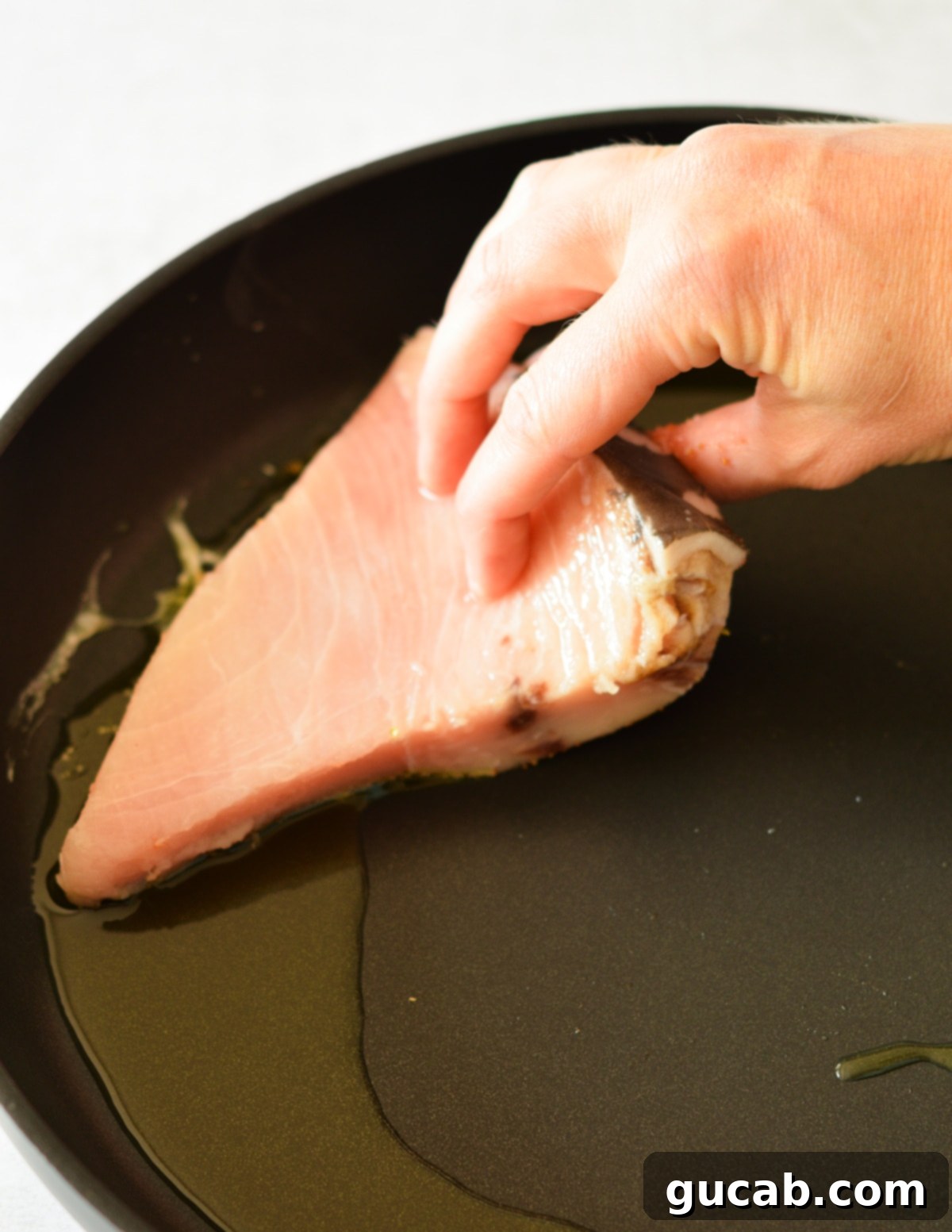 A hand carefully placing a seasoned swordfish steak, seasoned side down, into a sizzling hot skillet with olive oil.