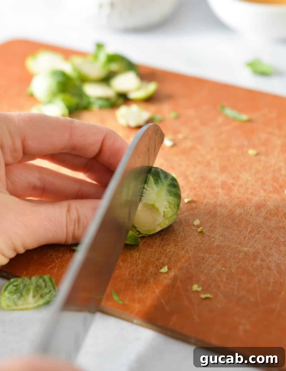 A chef's knife slicing a brussels sprout neatly in half lengthwise on a cutting board.