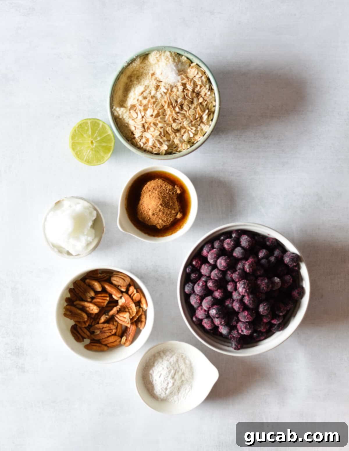 Arrangement of key healthy ingredients for blueberry crisp: almond flour, rolled oats, fresh lime, coconut oil, vibrant blueberries, and pecans.