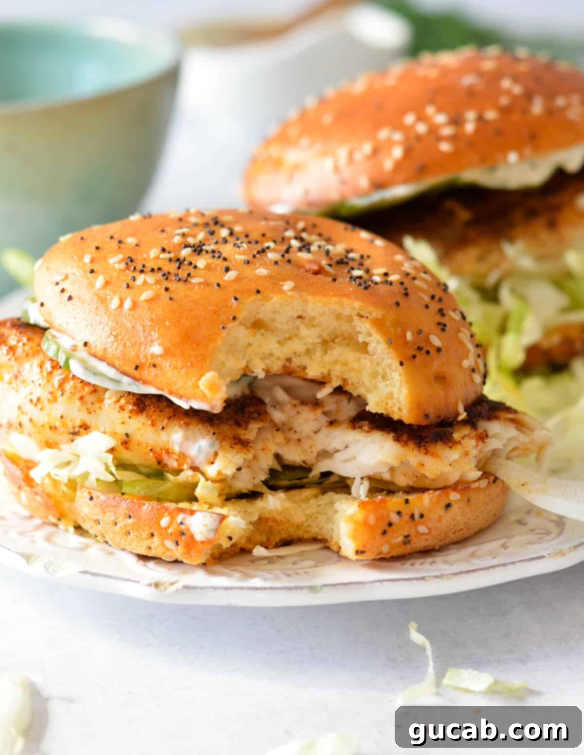 A close-up of a mahi mahi sandwich with a bite taken out, showing the layers of fish, lettuce, pickles, and aioli.