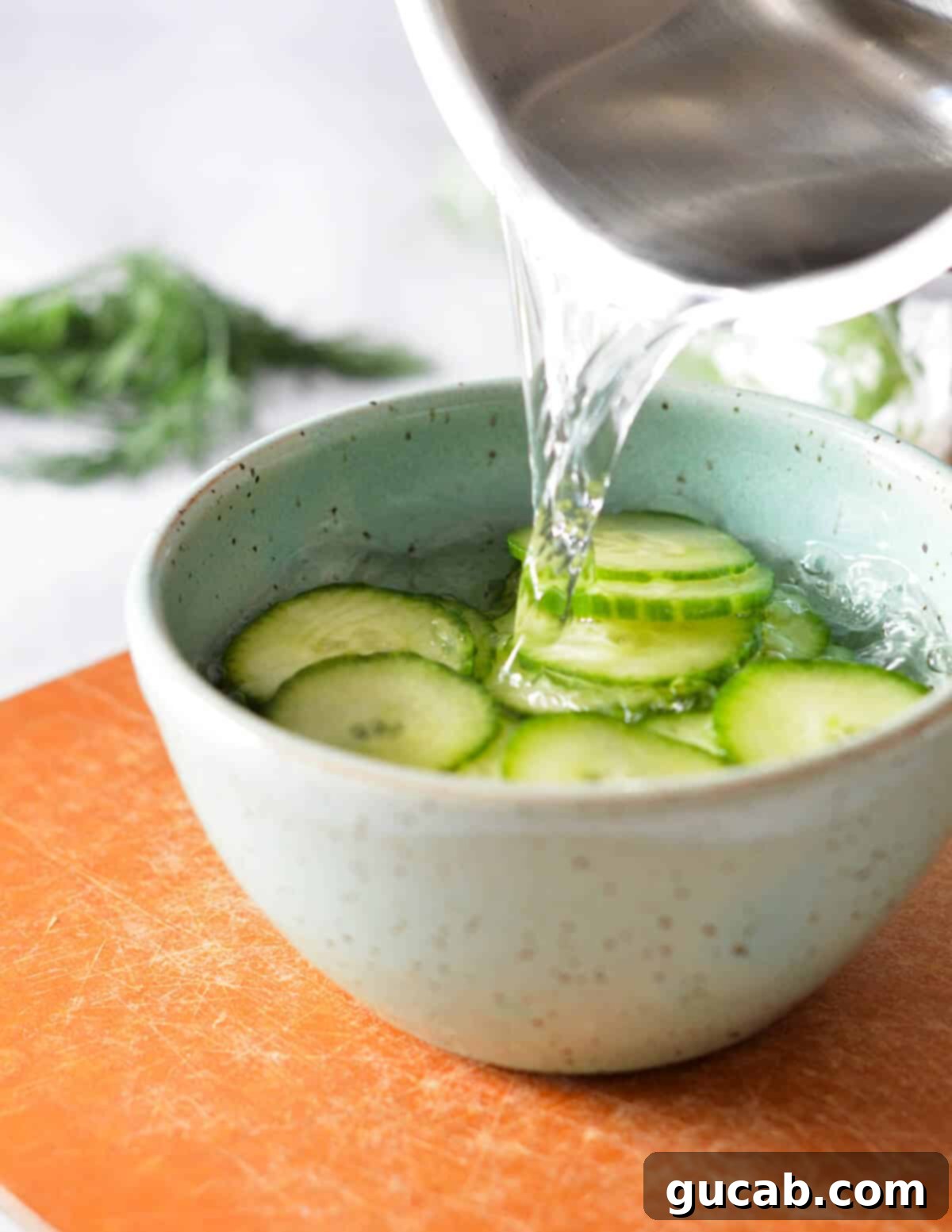 Hot vinegar mixture being poured over a bowl of thinly sliced cucumber rounds.