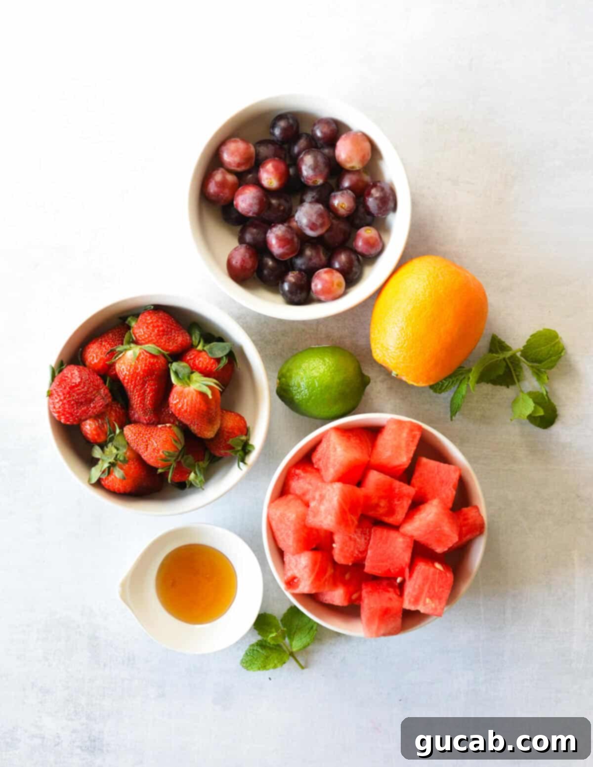 An artful arrangement of fresh ingredients including crisp grapes, vibrant strawberries, a navel orange, a lime, perfectly cubed watermelon, a jar of honey, and fresh mint leaves, all laid out on a clean white surface.