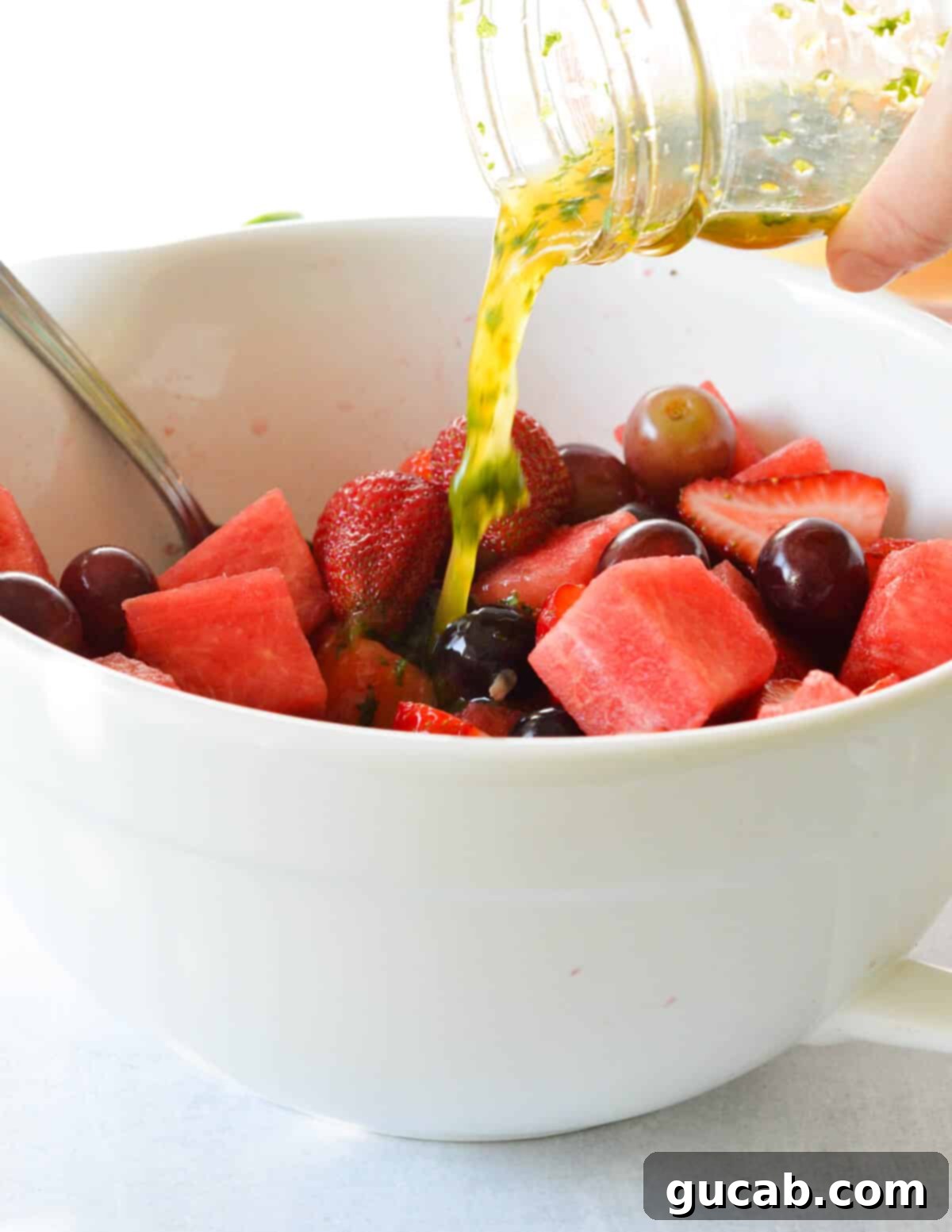 A stream of freshly prepared citrus dressing being poured over a large bowl of mixed fruit, including watermelon, grapes, and strawberries.