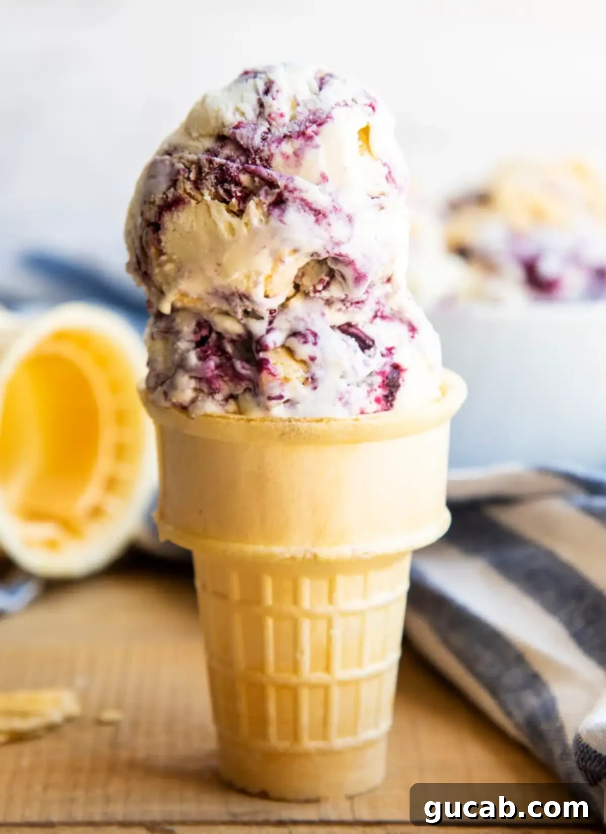 Two scoops of blueberry pie ice cream, flecked with blueberries and pie crust pieces, in a classic ice cream cone.