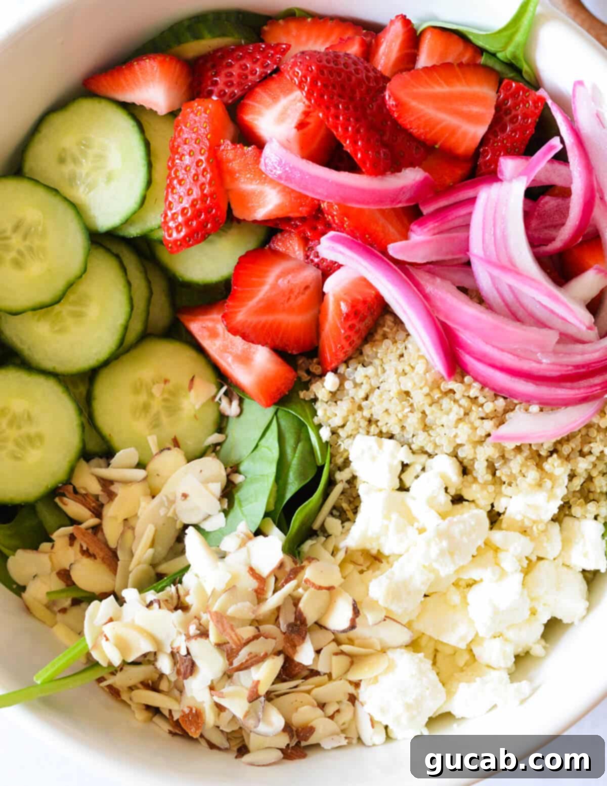 A medley of colorful and fresh salad toppings: sweet strawberries, crisp cucumbers, vibrant pickled red onions, crumbled feta cheese, crunchy sliced almonds, and wholesome quinoa, ready to be tossed.