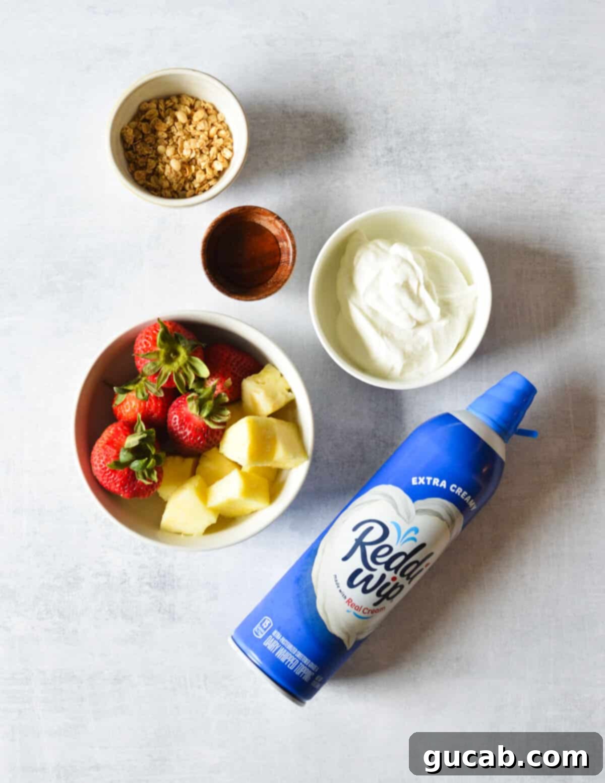 Essential ingredients for a yogurt parfait: Greek yogurt, fresh strawberries, diced pineapple, granola, honey, and whipped cream, arranged neatly.