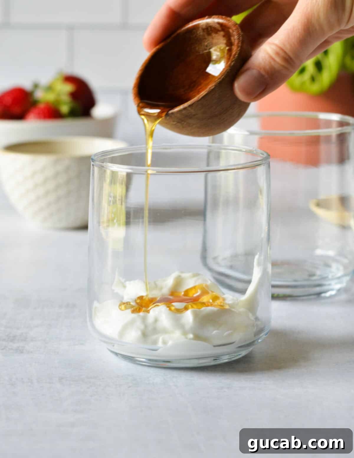 A steady stream of golden honey being drizzled over a layer of creamy Greek yogurt in a clear glass, ready for the next parfait layer.