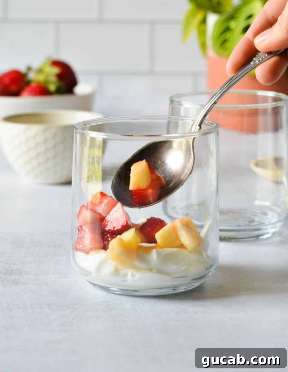 A spoon carefully adding a scoop of mixed diced fruit (strawberries and pineapple) into a glass over a layer of Greek yogurt and honey.