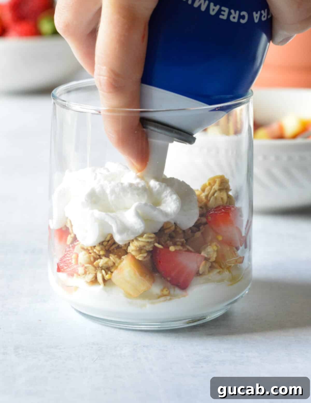 A can of whipped cream dispensing a fluffy white cloud onto a layer of fruit and granola in a glass, completing a parfait layer.