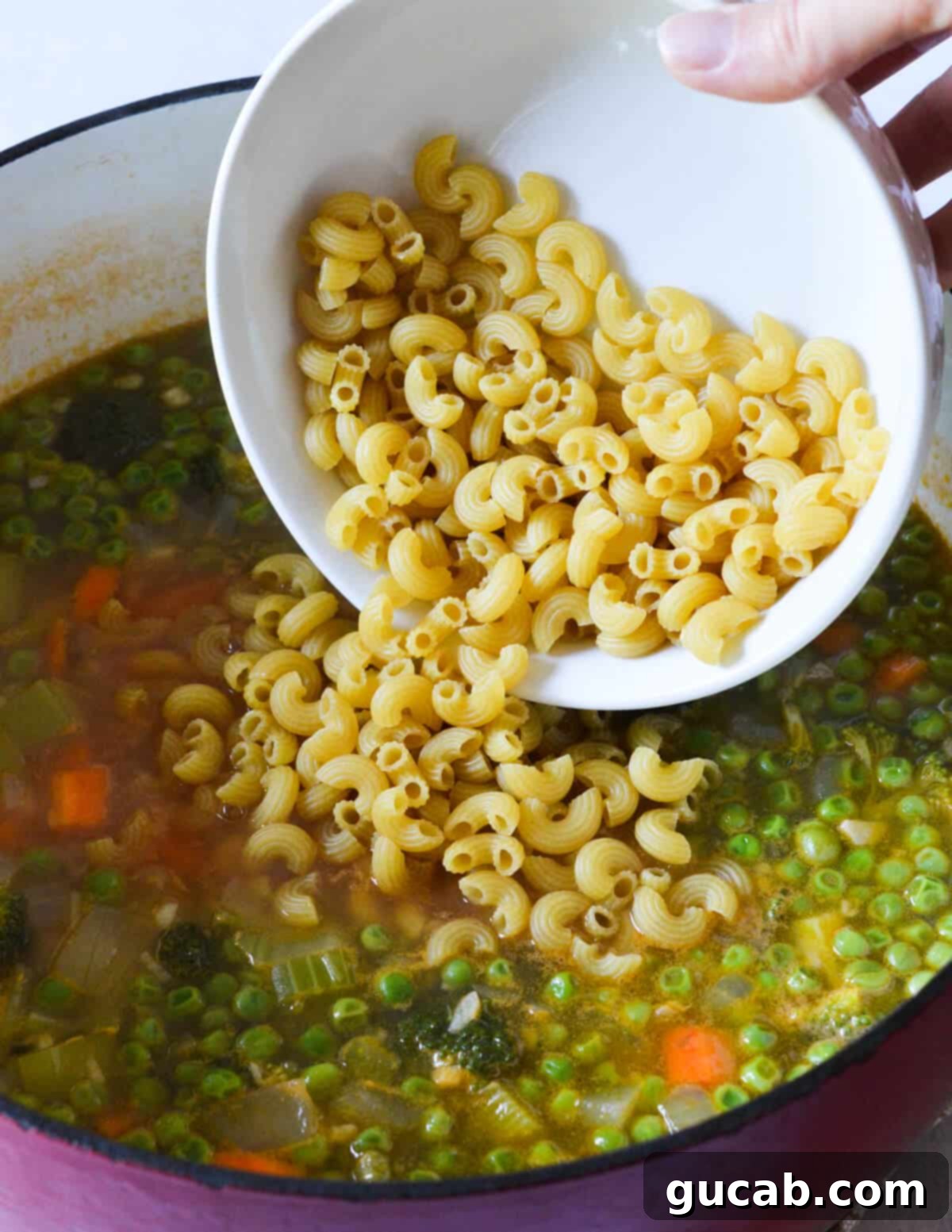 Vibrant Veggie Noodle Bowl 8 Uncooked elbow pasta being carefully added to a large pot of simmering vegetable soup, ready to cook directly in the flavorful broth.