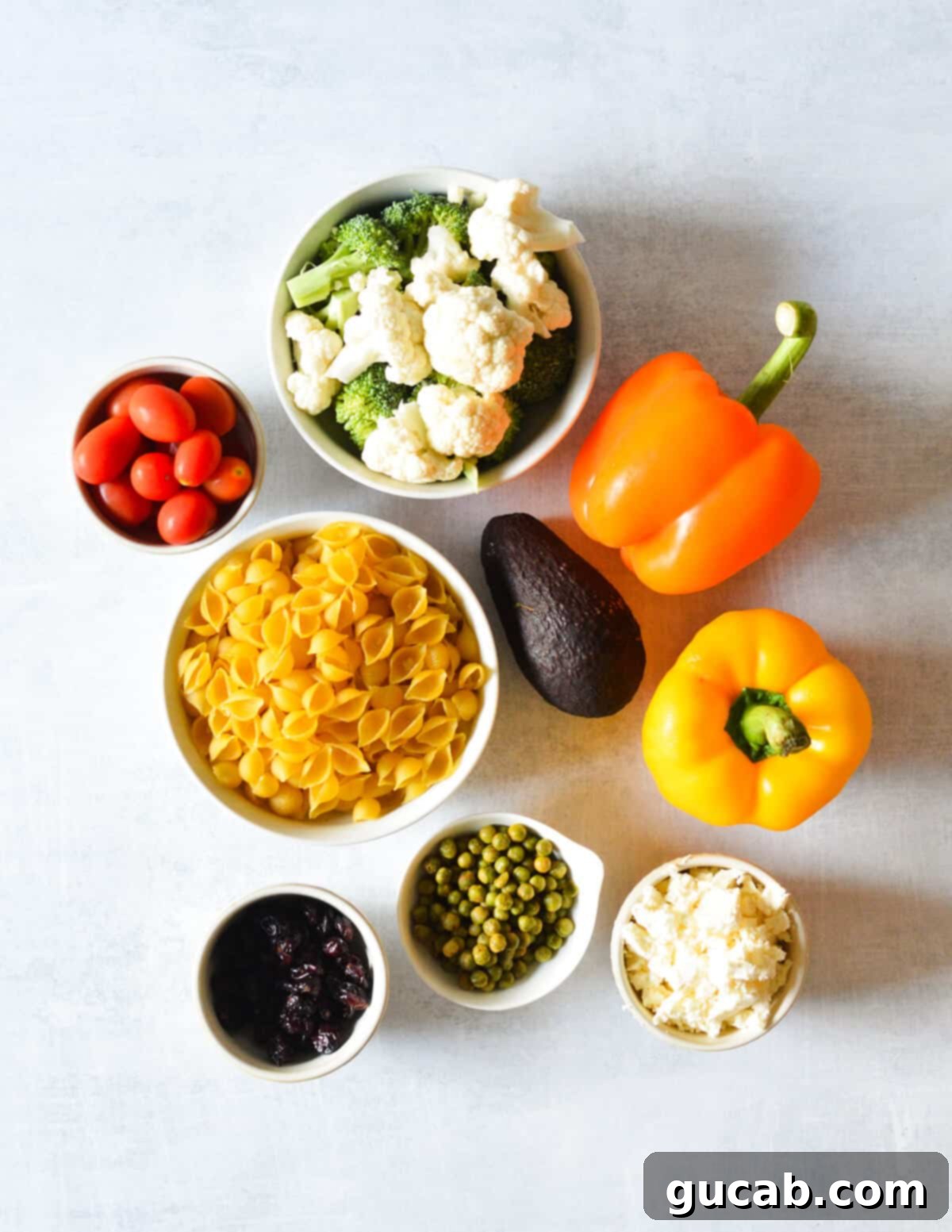 Dawn's Delightful Plant-Based Pasta Toss 3 A flat lay of fresh ingredients including uncooked shell pasta, broccoli florets, cauliflower florets, bell peppers, avocado, grape tomatoes, dried peas, dried cranberries, and crumbled feta cheese, ready for the pasta salad.